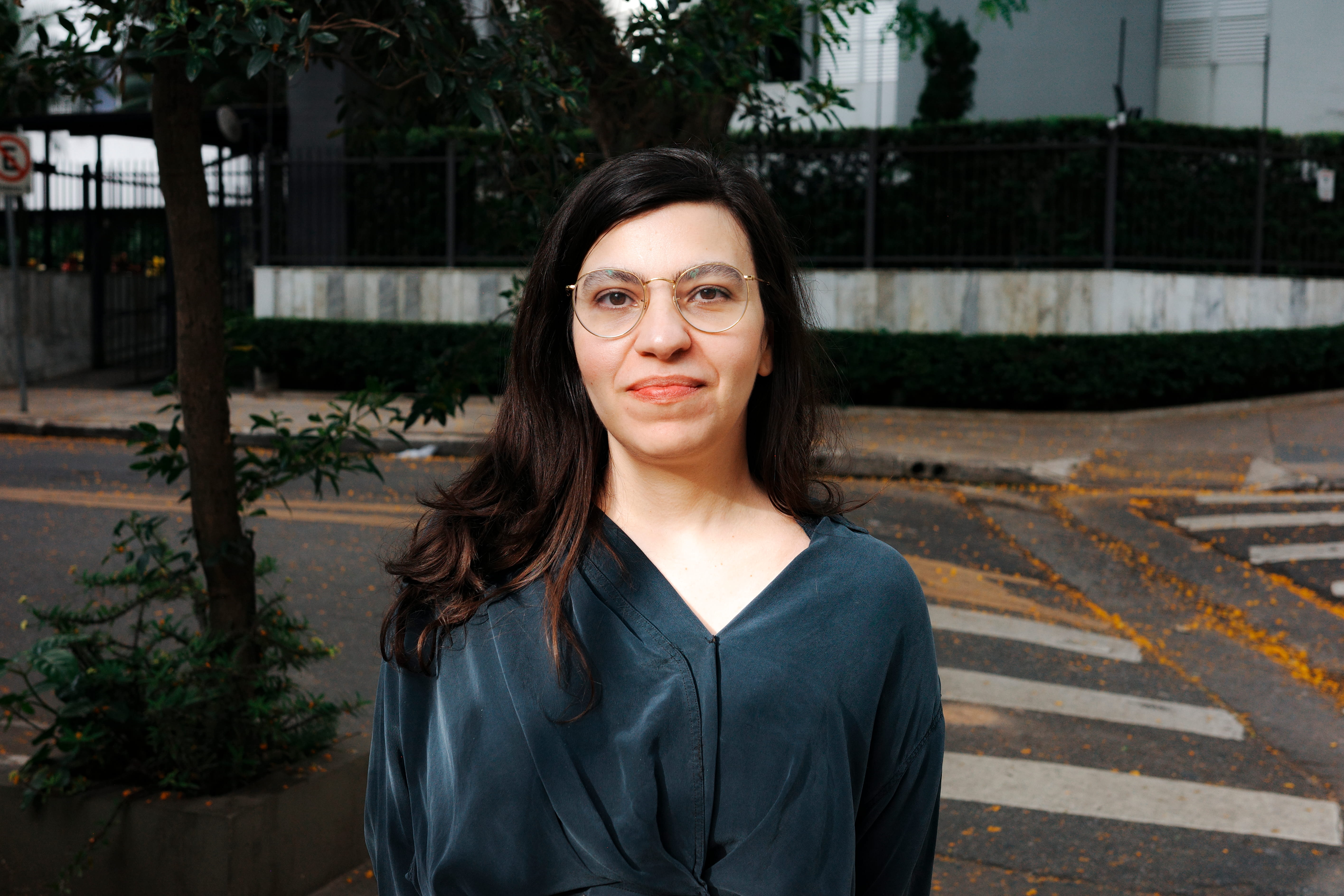 Foto de Nat&eacute;rcia Pontes, mulher branca, de cabelo castanho escuro longo, usa &oacute;culos e sorri para a c&acirc;mera, vestindo uma camisa azul marinho. 